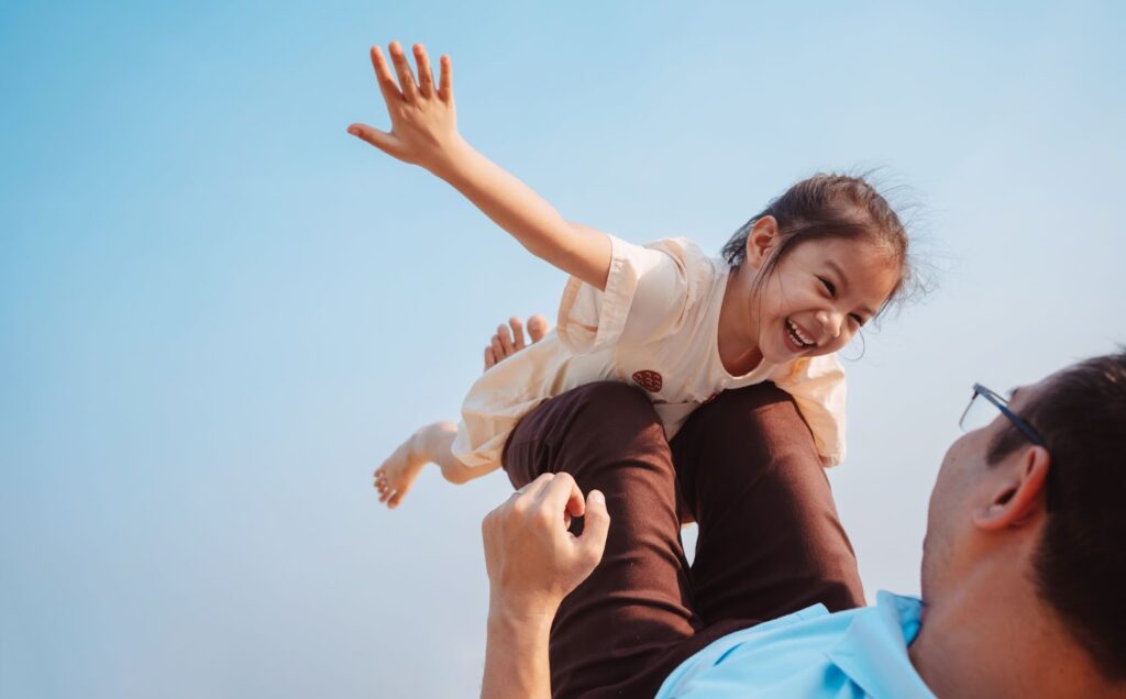 A child joyfully raised on an adult's shoulders against a clear blue sky.