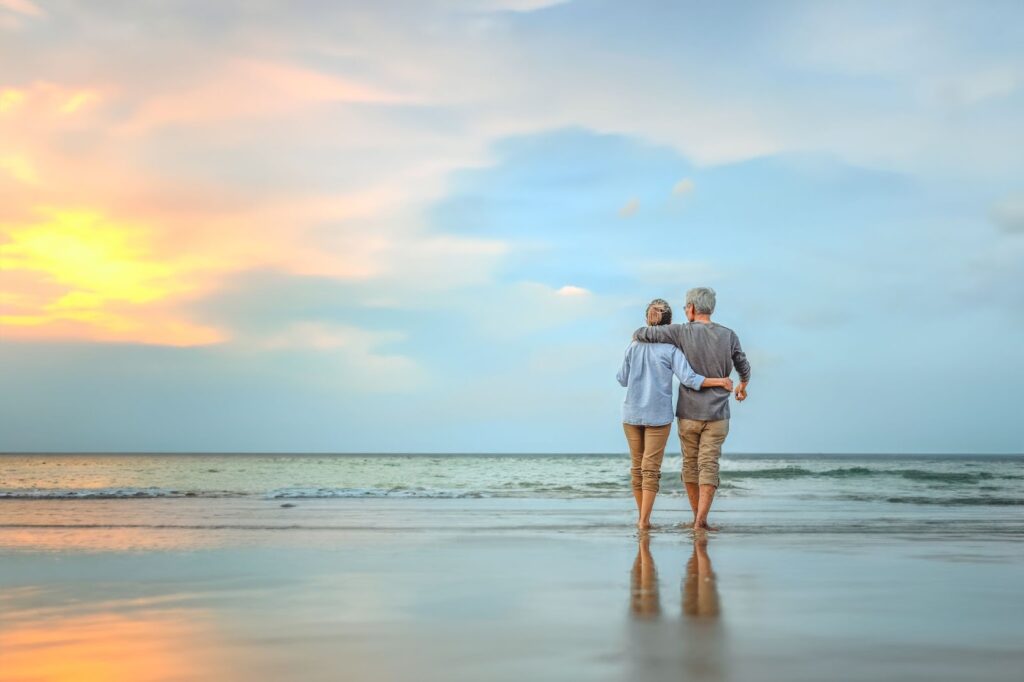 Elderly couple walking arm-in-arm on a beach at sunset, facing the ocean.